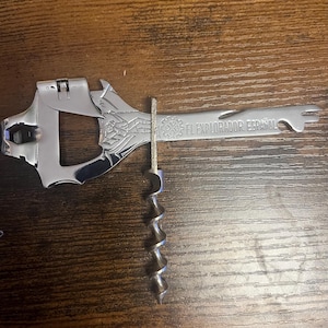 May include: A chrome-finished, multi-tool wine opener with a corkscrew and the text "EL EXPLORADOR ESPAÑOL". The opener has a wrench-like design with decorative details.