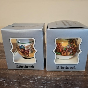May include: Two grey cardboard boxes with cut-out windows, each containing a round ceramic ornament. The left ornament features a child in a winter coat, while the right ornament displays a Christmas tree and gifts. Both boxes are labelled Alderbrook.