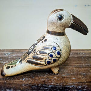 May include: A ceramic bird figurine with a cream-colored base, featuring hand-painted brown and blue floral and feather patterns. The bird has a prominent dark brown curved beak, a round black eye, and a smooth, rounded body shape.