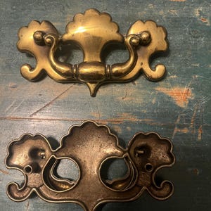 Peut inclure: Deux poignées de tiroir antiques en laiton. La poignée supérieure est en laiton massif avec un motif floral décoratif. La poignée inférieure a un design similaire, mais avec un dos creux et une finition vieillie plus foncée. Les deux ont une poignée incurvée.