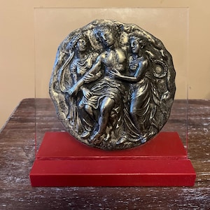 May include: A silver-toned relief sculpture depicting three figures in classical attire. The sculpture is mounted on a clear acrylic panel and a red rectangular base. The figures are in a close embrace, suggesting a narrative scene.