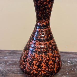 May include: A ceramic vase with a speckled orange and black pattern. The vase has a bulbous body and a slender neck, standing approximately 20 cm tall. The base is a dark brown colour.
