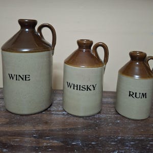 May include: Three ceramic jugs with brown tops and handles, labelled "WINE", "WHISKY", and "RUM" in black text. The jugs are beige and sit on a wooden surface. The jugs vary in size, with the "WINE" jug being the largest.