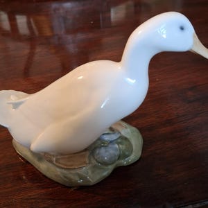 Peut inclure: Figurine de canard en céramique blanche avec un bec beige clair et des yeux bleus. Le canard est perché sur une base verte et beige. L'objet est décoratif.
