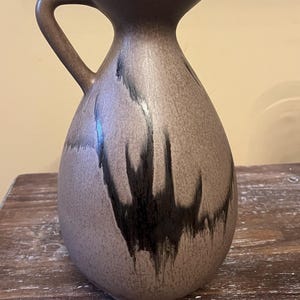 May include: A brown ceramic pitcher with a handle. The pitcher has a light brown base color with dark brown, dripped-style accents. The pitcher has a wide base that tapers to a narrow neck and a flared opening.