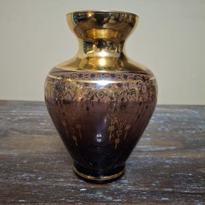 May include: A decorative vase with a gold rim and a gold interior. The vase has a dark purple body with gold floral designs. The base of the vase is black with a gold trim. The vase is on a wooden surface.
