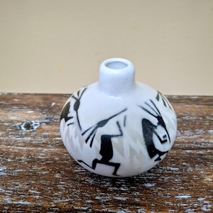 May include: A small, round, white ceramic vase with a narrow neck. The vase is decorated with black and grey stylized figures and geometric patterns. The vase is on a wooden surface.