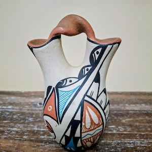 May include: A ceramic vessel with a double-spout design connected by a bridge handle. The off-white surface features hand-painted geometric patterns in black, terracotta orange, and light blue, creating a symmetrical and intricate visual display.