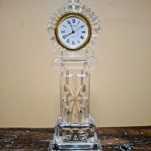 May include: A clear glass mantel clock featuring a faceted, geometric body and a circular clock face with a gold-toned bezel. The white dial displays black Roman numerals and thin black hands, with the text "WATERFORD" printed on the face.