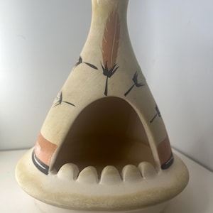 May include: Cream ceramic ashtray in a teepee shape. The ashtray has a wide base and a central opening. It is decorated with a brown feather and black floral accents. The rim has grooves for resting cigarettes.