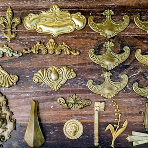 Peut inclure: Une collection de diverses pièces de quincaillerie de meubles en métal doré, comprenant des poignées de tiroir ornées, des écussons décoratifs, des motifs fleur de lys, un médaillon tête de lion et une tige florale sur bois sombre.