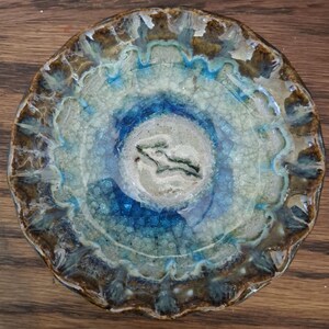 May include: A handmade ceramic bowl with a unique, organic design. The bowl features concentric circles of color, transitioning from a deep blue center to shades of turquoise, white, and brown around the edges. The bowl's surface has a crackled, textured appearance.