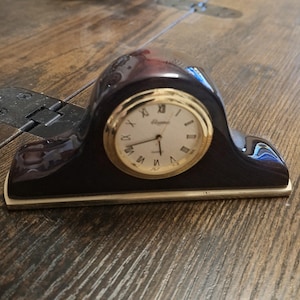 May include: A mantel clock with a dark mahogany-colored arched wooden frame and a gold-toned metal base. The clock face is white with black Roman numerals, gold-colored hands, and the word Quartz printed in black text below the center point.