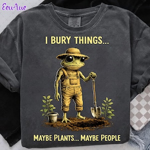 May include: Dark gray sweatshirt with a humorous graphic of a frog wearing overalls and a straw hat, holding a shovel. The text reads "I BURY THINGS... MAYBE PLANTS... MAYBE PEOPLE."