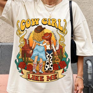 May include: A cream-colored t-shirt featuring a graphic print of two figures in cowboy hats standing in a desert landscape with cacti and red roses. The text COW GIRL and LIKE ME surrounds the illustration, with I KNEW IT I KNEW YOU in circles.