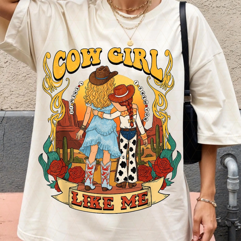 May include: A cream-colored t-shirt featuring a graphic print of two figures in cowboy hats standing in a desert landscape with cacti and red roses. The text COW GIRL and LIKE ME surrounds the illustration, with I KNEW IT I KNEW YOU in circles.