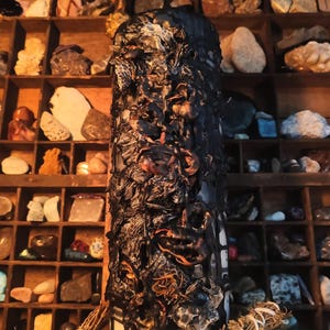 May include: A cylindrical pillar candle featuring a thick, textured black wax coating with intricate, organic, melted-like patterns. The candle is positioned upright, displaying a dark, uneven surface with visible layers and deep crevices throughout.
