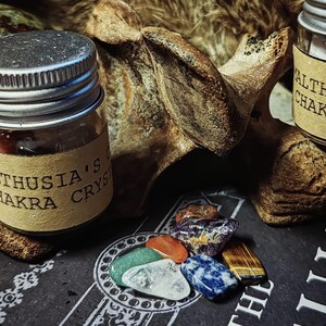 May include: A small glass jar with a silver lid filled with colourful crystals. The jar has a label that reads "Thusia's Chakra Crystal". There are several colourful crystals scattered on a black surface.