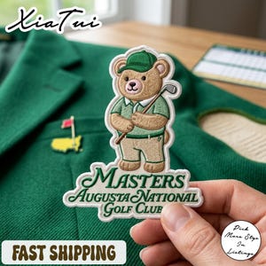 Op de afbeelding: Geborduurde patch met een beer die een groene pet en shirt draagt en een golfclub vasthoudt. De patch leest "MASTERS AUGUSTA NATIONAL GOLF CLUB" in groen en wit. Een kleine pin met een vlag is ook zichtbaar. De achtergrond is een groene jas.