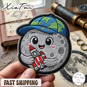 May include: Embroidered patch featuring a smiling moon wearing a blue and green Earth-themed cap and holding a red and white rocket. The patch has a gray border and is on a light background. The text "FAST SHIPPING" is at the bottom.