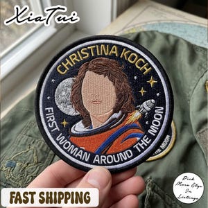 以下が含まれることがあります： クリスティーナ・コックを描いた刺繍パッチ。テキストは「First Woman Around The Moon」。デザインには肖像画、ロケット、星、月がダークブルーの背景に描かれており、黒い縁取りがあります。