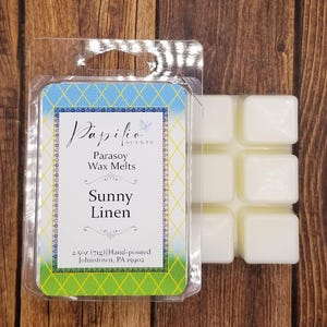 May include: A package of Sunny Linen scented Parasoy wax melts. The wax melts are white and in a square shape. The packaging has a blue and green design with the text "Papilio Scents" and "Sunny Linen".