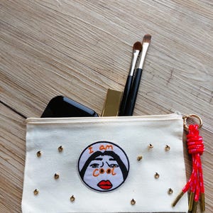 Könnte beinhalten: Eine weiße Make-up-Tasche aus Canvas mit goldenen Nieten und einem Reißverschluss. Die Tasche hat einen gestickten Aufnäher mit den Worten "I am cool" und einem Cartoon-Gesicht. Make-up-Pinsel und Lippenstift sind darin.