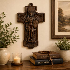 May include: A dark brown wooden wall cross featuring a relief carving of Saint Michael the Archangel with wings, holding a sword and shield, standing over a dragon. The cross has a decorative border and a small hole at the top for hanging.