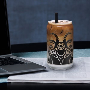 May include: A glass with a black and white graphic of a goat with wings and a star on its head. The glass is filled with iced coffee.