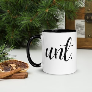 May include: White ceramic mug with a black rim and handle. The mug has the word "unit" written in black cursive on the front.