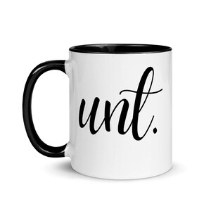 May include: White ceramic mug with a black rim and handle. The mug has the word "aunt" written in black cursive script.