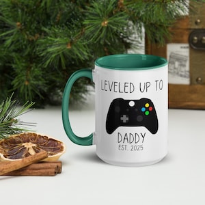 May include: White ceramic mug with a green rim. The mug features a black and white illustration of a video game controller and the text "Leveled Up To Daddy Est. 2025".