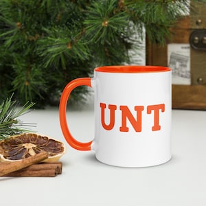 Peut inclure: Une tasse en céramique blanche avec un bord et une poignée orange. La tasse a le mot "UNT" imprimé en orange sur le devant.