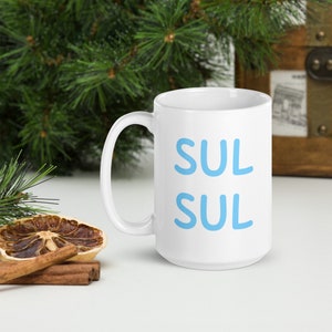 May include: White ceramic mug with a blue inscription that reads "Sul Sul" in a playful, handwritten style.