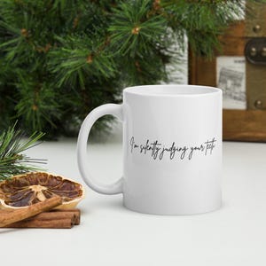 May include: White ceramic mug with a curved handle. The mug features the phrase "I'm silently judging your teeth" in black script. The mug is on a white surface with a dried orange slice and cinnamon sticks.