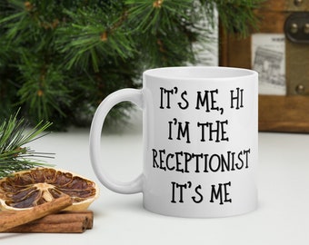 May include: White ceramic mug with the text "It's me, Hi I'm the Receptionist It's Me" in black lettering. The mug has a curved handle and is set against a background of greenery and a wooden chest.
