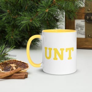 May include: White ceramic mug with a yellow rim and handle. The mug has the word "UNT" printed in yellow on the front.