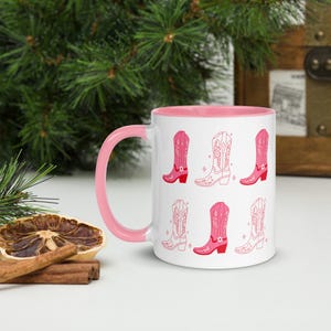 Peut inclure: Une tasse en céramique blanche avec un bord et une anse roses. La tasse présente un motif répété de bottes de cow-boy roses avec des contours blancs.