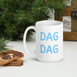 May include: White ceramic coffee mug with a blue inscription that reads "DAG DAG".