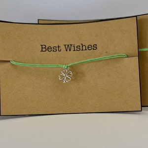 May include: A green string bracelet with a silver four-leaf clover charm. The bracelet is displayed on a brown card with the text "Best Wishes".