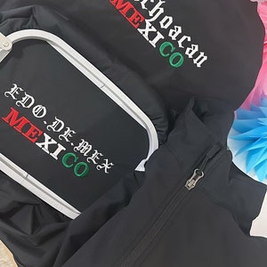 May include: Black embroidered jacket with a zipper and a green, white, and red embroidered Mexican coat of arms on the right side. The jacket also has the words "Michoacan MEXICO" and "EDO DE MEX MEXICO" embroidered on it.
