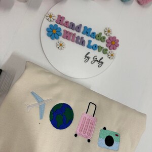 K&ouml;nnte beinhalten: Ein beiger Pullover mit gestickten Bildern eines Flugzeugs, eines Globus, eines rosafarbenen Koffers und einer Kamera. Der Text "Hand Made With Love by Gaby" befindet sich auf einem wei&szlig;en Kreis mit bunten Blumen.