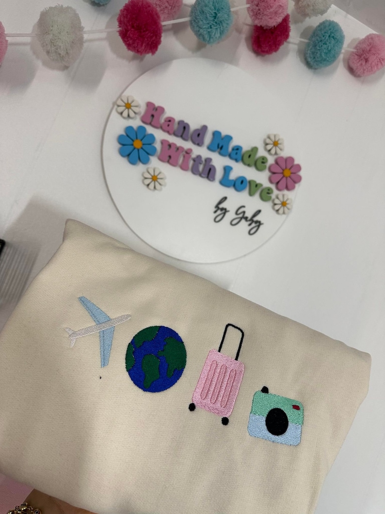 K&ouml;nnte beinhalten: Ein beiger Pullover mit gestickten Bildern eines Flugzeugs, eines Globus, eines rosafarbenen Koffers und einer Kamera. Der Text "Hand Made With Love by Gaby" befindet sich auf einem wei&szlig;en Kreis mit bunten Blumen.