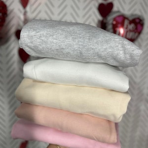 K&ouml;nnte beinhalten: Ein Stapel von f&uuml;nf gefalteten Sweatshirts in verschiedenen Farben: grau, wei&szlig;, beige, rosa und hellrosa.
