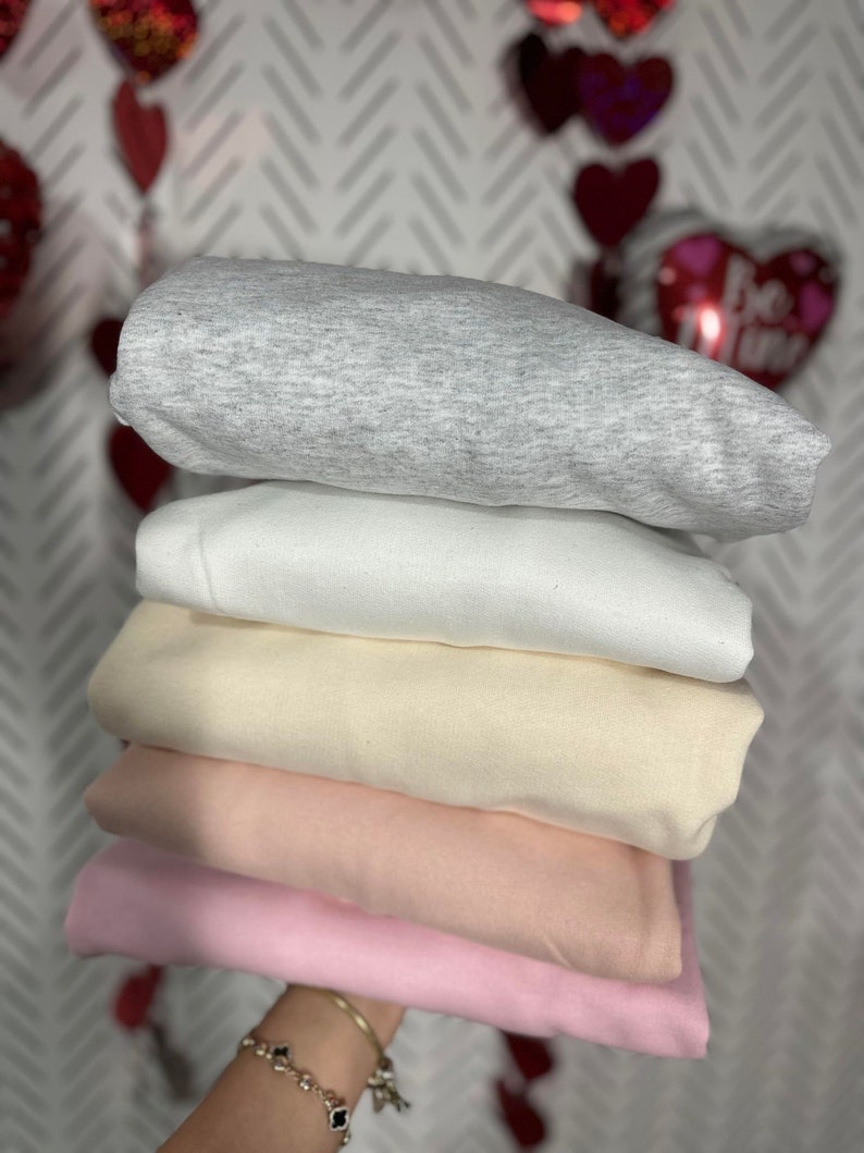 K&ouml;nnte beinhalten: Ein Stapel von f&uuml;nf gefalteten Sweatshirts in verschiedenen Farben: grau, wei&szlig;, beige, rosa und hellrosa.