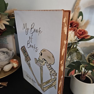May include: A hardback book titled "My Book of Books" with a skeleton illustration on the cover. The spine is brown with a decorative pattern. The book is on a black surface.