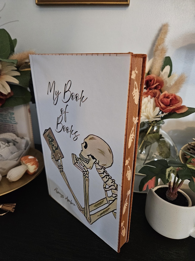 May include: A hardback book titled "My Book of Books" with a skeleton illustration on the cover. The spine is brown with a decorative pattern. The book is on a black surface.