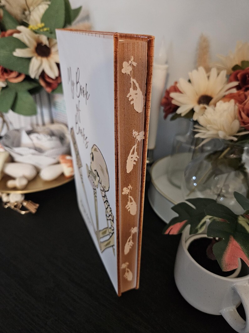 May include: A hardback book titled "My Book of Bones" with a white cover and an orange spine. The spine has a repeating floral pattern. The cover features a skeleton illustration and handwritten text.
