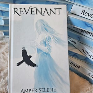 Könnte beinhalten: Ein gebundenes Buch mit dem Titel REVENANT von Amber Selene. Das weiße Cover zeigt eine blau getönte Illustration einer geisterhaften Figur in einem fließenden Kleid und einen fliegenden schwarzen Vogel. Der Titel steht oben in Schwarz.