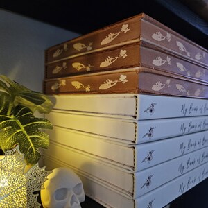 May include: A collection of books with the title "My Book of" on the spines. The top books are brown with a decorative design, while the lower books are white with a floral accent. A small skull and decorative leaf are in the foreground.